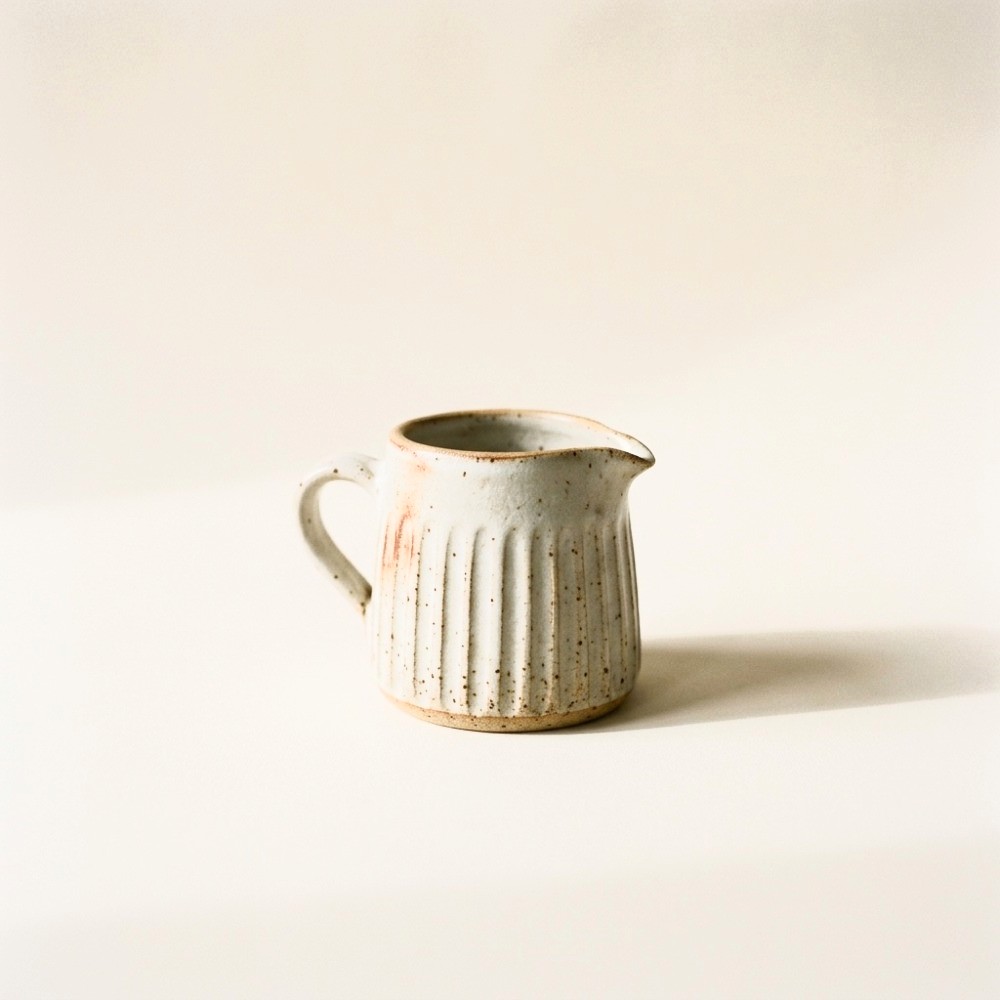 A small fluted stoneware milk jug with a pinched pouring spout and a matte cream glaze over speckled clay.