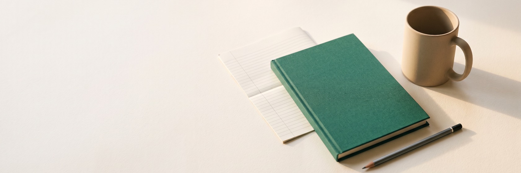 A green cloth-bound ledger book closed on a pale desk beside a ruled notepad, a plain stoneware mug and a pencil, in low afternoon light.
