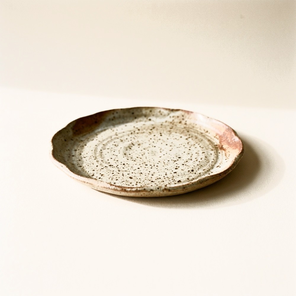 A stoneware side plate with an oatmeal glaze, iron flecks through the surface and a slightly undulating rim.